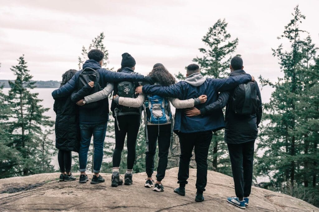 Group of hikers with backpacks standing arm in arm on a rocky overlook surrounded by forest and water
